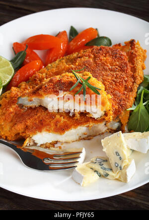 Lecker gebratene Panierte weißen Fisch steak Schneiden in zwei Hälften auf weiße Platte mit glasierten Karotten, Kalk, Scheiben, blue Schimmelkäse und Rucola Salat serviert, ing Stockfoto