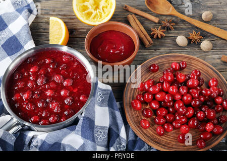 Lecker cranberry Sauce in Topf und Schüssel auf hölzernen Tisch mit frischen Cranberry auf Platte und Zutaten auf Hintergrund, authentische Rezept, horiz. Stockfoto