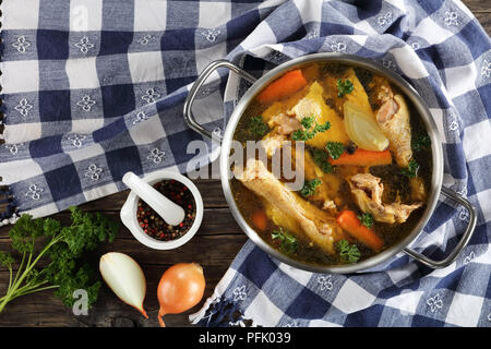 Klare Hühnerbrühe mit Stücken von Henne und Hahn Fleisch auf die Knochen in einem Metall Kochtopf auf Holztisch mit Küchentuch und Zutaten auf Holz- ta Stockfoto