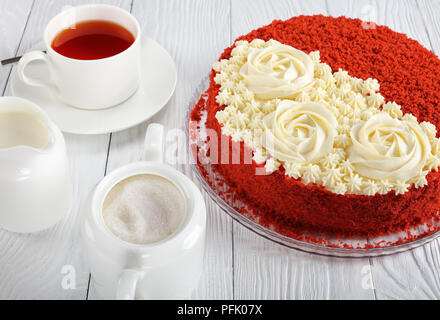 Leckere hausgemachte Red Velvet Cake gekrönt mit schön cremig Rosen auf Glas Teller am Tisch bei einer Tasse Tee serviert, Sugar Bowl und kanne Milch, Anzeigen Stockfoto