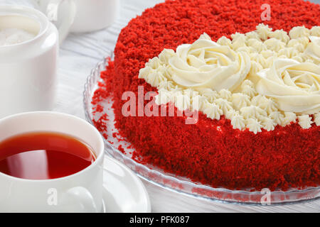 In der Nähe von köstlichen hausgemachten Red Velvet Cake gekrönt mit schönen cremefarbenen Rosen und sprengte mit roter Schwamm Kuchen Krümel auf Glas Schale mit Tasse Stockfoto