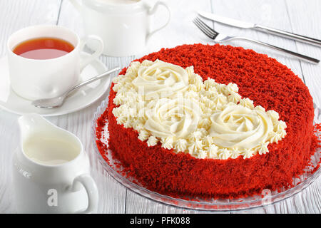 Leckere hausgemachte Red Velvet Cake gekrönt mit schön cremig Rosen auf Glas Teller am Tisch bei einer Tasse Tee serviert, Sugar Bowl und kanne Milch, Seite Stockfoto