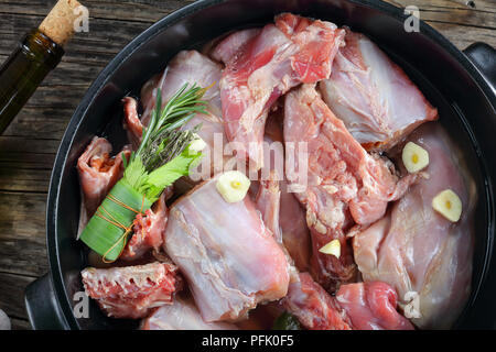 Stück Kaninchen roh mariniert mit Weißwein, Knoblauch, Gewürze und Bündel von aromatischen Kräutern oder bouquet garni in schwarz Auflaufform auf dunklen Holztisch, Stockfoto