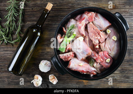 Stück Kaninchen roh mariniert mit Weißwein, Knoblauch und Bündel von aromatischen Kräutern oder bouquet garni in schwarz Auflaufform auf Tisch mit einer Flasche Wein Stockfoto