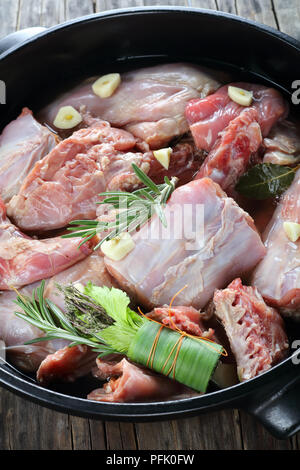 Stück Kaninchen roh mariniert mit Weißwein, Knoblauch, Gewürze und Bündel von aromatischen Kräutern oder bouquet garni in Auflaufform auf alten, dunklen Holzbrettern, Stockfoto