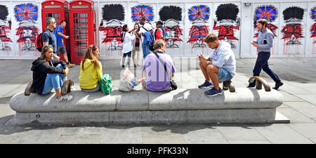 Touristen in St Martin's Place, London, England, UK. Traditionelle rote Telefonzellen und Graffiti Wachposten durch Künstler Nathan Bowendecorating constructi Stockfoto