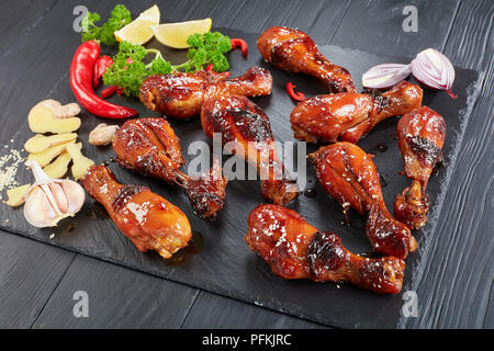 Lecker gegrilltes knusprigen saftigen teriyaki Chicken Drumsticks bestreut mit Sesam auf schwarzen Stein Fach auf Holztisch mit Petersilie, Knoblauch und Zitrone Stockfoto