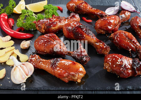 Close-up lecker Gegrilltes knusprigen saftigen teriyaki Chicken Drumsticks bestreut mit Sesam auf schwarzen Stein Fach auf Holztisch mit Petersilie, garli Stockfoto