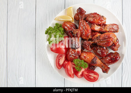 Lecker gegrilltes knusprigen saftigen teriyaki Chicken Drumsticks bestreut mit Sesam auf weiße Platte mit Tomaten, Petersilie und Zitronenscheiben, die Aussicht von ein Stockfoto