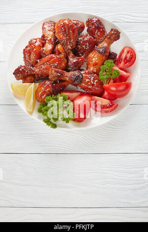 Lecker gegrilltes knusprigen saftigen teriyaki Chicken Drumsticks bestreut mit Sesam auf weiße Platte mit Tomaten, Petersilie und Zitronenscheiben, vertikale vi. Stockfoto