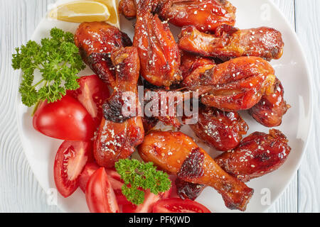 Gegrillte knusprigen saftigen teriyaki Chicken Drumsticks bestreut mit Sesam auf weiße Platte mit Tomaten, Petersilie und Zitronenscheiben, von oben betrachten, Stockfoto