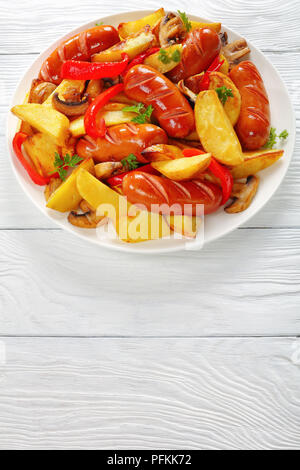 Hot köstliche gegrillte Würstchen mit Bratkartoffeln Keile, Champignons und Petersilie auf einer weißen Platte auf Holztisch, vertikale Ansicht von oben Stockfoto