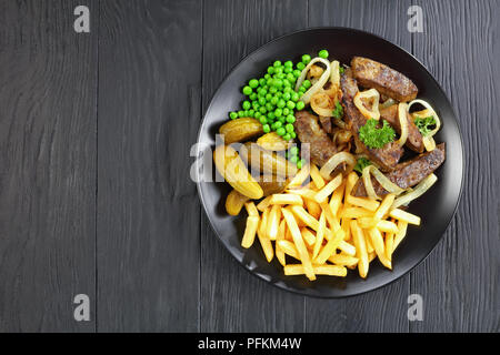 Lecker gebratene Kalbsleber mit Pommes, eingelegte Gurken, grüne Erbsen auf schwarzem Teller auf Holz- Tabelle, Ansicht von oben Stockfoto