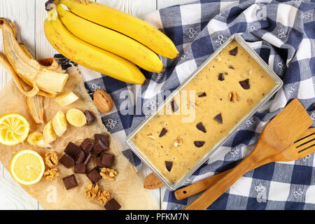 Nicht gegarte Banane Brot mit Nüssen und Schokolade Stücke in eine Kastenform auf Holztisch mit Küchentuch, Spatel und Zutaten auf Hintergrund, Anzeigen Stockfoto