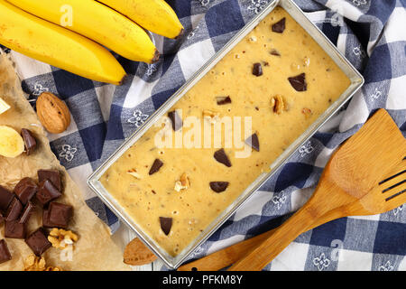 Raw Banane Brot mit Nüssen und Schokolade Stücke in eine Kastenform auf Holztisch mit Küchentuch, Spatel und Zutaten auf Hintergrund, Ansicht von Stockfoto