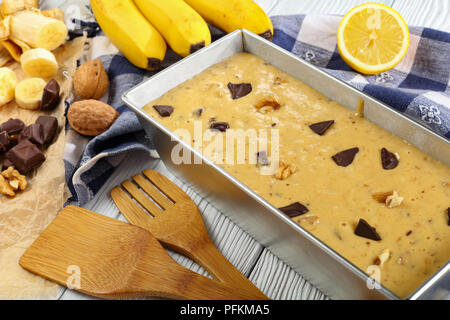 Close-up ungekochten Banane Brot mit Nüssen und Schokolade Stücke in eine Kastenform Vorbereitung zum Backen. Küchentuch, Spatel und Zutaten auf Bac Stockfoto