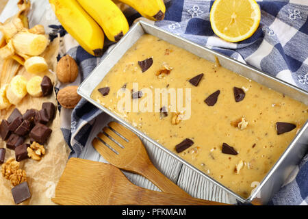 Close-up ungekochten Banane Brot mit Nüssen und Schokolade Stücke in eine Kastenform auf Holztisch mit Küchentuch, Spatel und Zutaten auf zurück Stockfoto