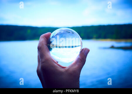 Hand mit einem Glas Kugel spiegelt den See und den Wald. Konzept der Umwelt, Naturschutz, Ökologie. Selektiver Fokus Stockfoto