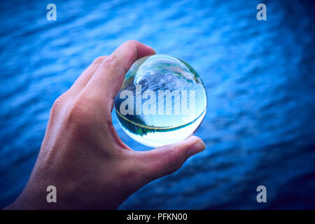 Hand mit einem Glas Kugel spiegelt den See und den Wald. Konzept der Umwelt, Naturschutz, Ökologie. Selektiver Fokus Stockfoto