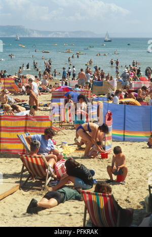 England, Dorset, Weymouth, crowded beach with people sunbathing on deck chairs and swimming in the sea. Stockfoto