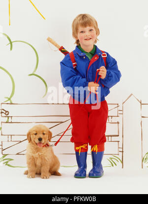 Blonde junge tragen Gummistiefel und mit Rucksack auf dem Rücken mit Schirm kleben, ständigen Holding führen zu Halsband von Golden Retriever Welpe, Abbildung: Garden Gate im Hintergrund, Vorderansicht. Stockfoto
