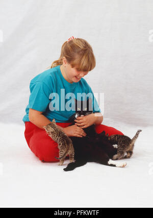 Lächelnde blonde Mädchen sitzen auf dem Boden spielen mit schwarzen Katze (Felis silvestris catus) und Wurf von schwarzen und tabby Kätzchen. Stockfoto
