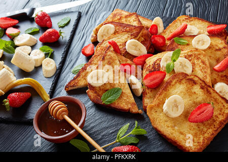 French Toast mit appetitlich goldbraune Kruste und flauschige Innenseite, serviert auf einem Schiefer mit frischen Erdbeeren, Bananenscheiben, Minze und Honig. ing Stockfoto