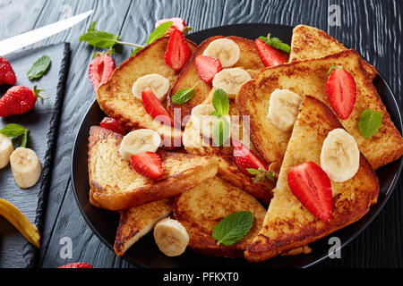 French Toast mit appetitlich goldbraune Kruste und flauschige Innenseite serviert auf einem Teller mit frischen Erdbeeren, Bananenscheiben, Minze. Zutaten auf Schnitt Stockfoto