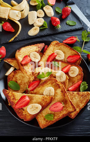 French Toast mit appetitlich goldbraune Kruste und flauschige Innenseite auf schwarzem Teller serviert mit frischen Erdbeeren, Banane, Minze. Zutaten auf Stockfoto