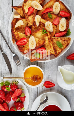 French Toast mit appetitlich goldbraune Kruste und flauschige Innenseite auf Platte mit frischen Erdbeeren, Banane, Minze, Ahornsirup, Kaffee und Stockfoto