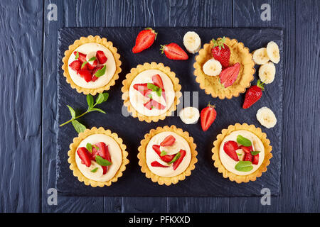 Törtchen mit Erdbeeren, Bananenscheiben mit Schlagsahne puddingpulver Sahne beladen - Diplomat auf schwarzem Schiefer Platte, Ansicht von oben, close-up Stockfoto