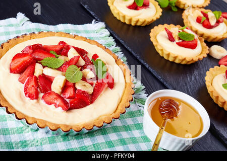 Leckere Törtchen mit Erdbeeren, Bananenscheiben mit Vanillesauce Creme auf einem schwarzen Schiefer Platte geladen. Start in das Backblech auf Textil- mat, Ansicht von eine Stockfoto