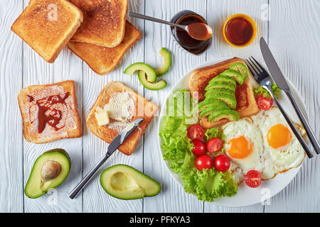 Gesundes Frühstück - Spiegeleier, frische Salate, Toasts mit Hefe ausbreiten und in Scheiben geschnittenen Avocado auf einer weißen Platte auf einem Holztisch, Ansicht von oben Stockfoto