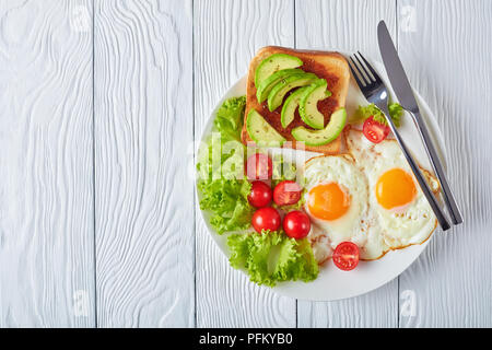 Gesunde australischen Frühstück - Spiegeleier, frischen Salat, Toast mit Hefe ausbreiten und in Scheiben geschnittenen Avocado auf einer weißen Platte auf einem Holztisch, Ansicht von oben Stockfoto