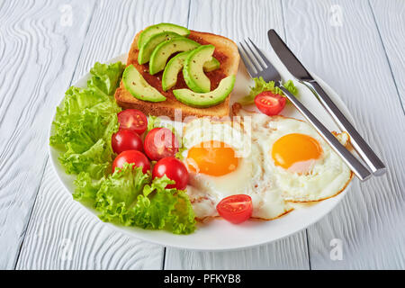 Englisches Frühstück - Spiegeleier, frischen Salat, Toast mit Hefe ausbreiten und in Scheiben geschnittenen Avocado auf einer weißen Platte auf einem Holztisch, horizontale Ansicht von oben Stockfoto