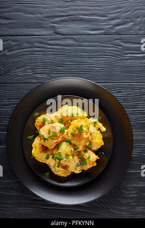 Neue Kartoffeln im Ofen gebacken mit Cheddar Käse butter Soße, auf schwarzen Platten auf einem schwarzen Holztisch serviert, vertikale Ansicht von oben Stockfoto