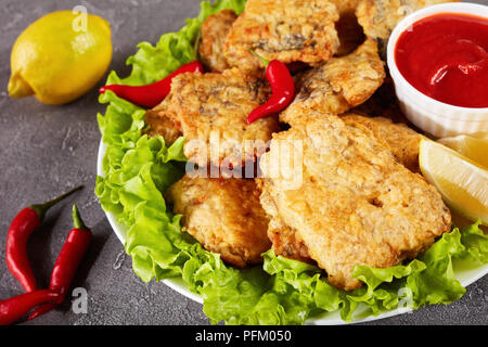 Gebratene Stücke von Seehecht auf einer weißen Platte auf einer konkreten Tabelle mit Zitronenscheiben, Tomatensauce und Paprika, Ansicht von oben, close-up Stockfoto