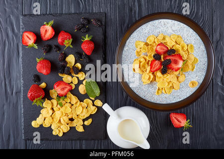 Gesundes Frühstück - Corn Flakes mit Chia Samen Pudding, frische Erdbeeren und Maulbeeren in einer Schüssel auf schwarzer Holztisch mit Zutaten auf Schnitt b Stockfoto