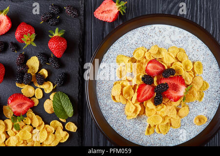 Gesundes Frühstück - Corn Flakes mit Chia Samen Pudding, frische Erdbeeren und Maulbeeren in einer Schüssel auf schwarzer Holztisch mit Zutaten auf Schnitt b Stockfoto