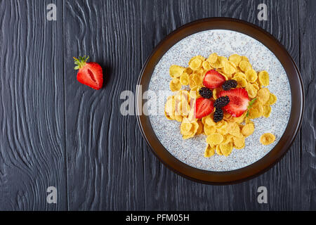 Teil von Chia Samen pudding gekrönt mit Cornflakes, frisch Erdbeeren, und Maulbeeren in einer Schüssel auf schwarzem Holz- Tabelle, Ansicht von oben, Nahaufnahme, Fla Stockfoto