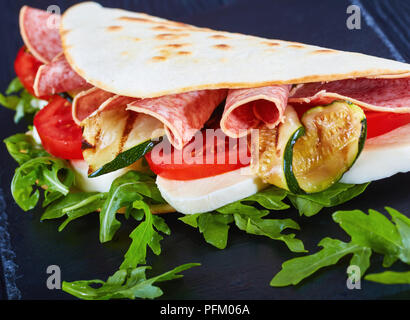 Close-up Italienisch sandwich Wraps oder Piadina mit Mozzarella, Tomaten, Salami Scheiben, gegrillten Zucchini und Rucola auf schwarzem Schiefer Platte, Ansicht von eine Stockfoto