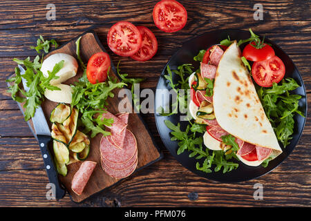 Frisch gebackene italienische Piadina mit Mozzarella, Tomaten, Salami Scheiben, gegrillten Zucchini und Rucola auf eine schwarze Platte mit Zutaten auf einem Holztisch Stockfoto