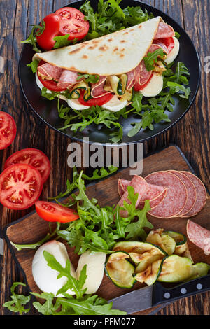 Iitalian Sandwich - piadina mit Mozzarella, Tomaten, Salami Scheiben, gegrillten Zucchini und Rucola auf eine schwarze Platte mit Zutaten auf einem Holztisch, Stockfoto