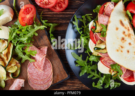 Frisch gebackene italienische Piadina mit Mozzarella, Tomaten, Salami Scheiben, gegrillten Zucchini und Rucola auf eine schwarze Platte mit Zutaten auf einem Holztisch Stockfoto