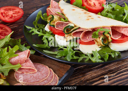 Sandwich - Italienisches piadina mit Mozzarella, Tomaten, Salami Scheiben, gegrillten Zucchini und Rucola auf eine schwarze Platte mit Zutaten auf einem Schneiden bo Stockfoto