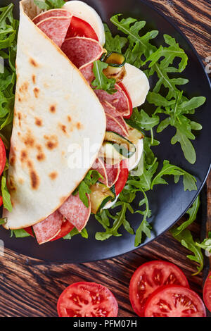 Sandwich - Italienisches piadina mit Mozzarella, Tomaten, Salami Scheiben, gegrillten Zucchini und Rucola auf eine schwarze Platte mit Zutaten auf einem Schneiden bo Stockfoto