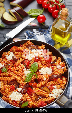 Vegetarische italienische Pasta Fusilli alla Norma mit Auberginen, Tomaten, Basilikum, ricotta Käse in einer Pfanne mit Zutaten im Hintergrund, vertikale v Stockfoto