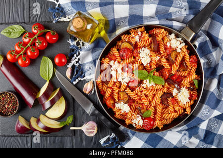 Vegetarische italienische Pasta Fusilli alla Norma mit Auberginen, Tomaten, Basilikum, ricotta Käse in einer Pfanne auf einem Holztisch mit Zutaten auf der Rückseite Stockfoto