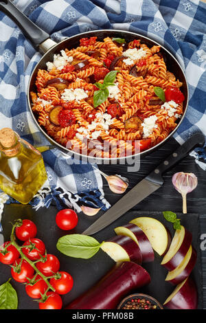 Vegetarische italienische Pasta Fusilli alla Norma mit Auberginen, Tomaten, Basilikum, ricotta Käse in einer Pfanne auf einem Holztisch mit Zutaten auf der Rückseite Stockfoto