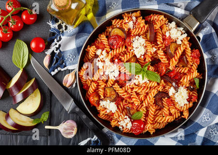 Italienische Pasta Fusilli alla Norma mit Auberginen, Tomaten, Basilikum, ricotta Käse in einer Pfanne auf einem Holztisch mit Zutaten im Hintergrund, ver Stockfoto